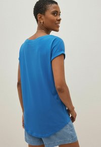 Blå kortärmad topp i mjukt material med rundad halsringning och något böjd nederkant. Bärs med ljusa jeansshorts.