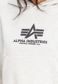Felpa grigia realizzata in tessuto morbido, con tasca frontale e un logo nero con ali e la scritta "ALPHA INDUSTRIES, KNOXVILLE, TENNESSEE, U.S.A."