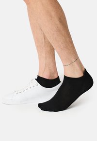 Schwarze Knöchelsocke neben einem weißen Sneaker. Die Socke hat eine glatte Textur und einen elastischen Bund. Der Sneaker hat ein Schnürdesign mit einer flachen Sohle.