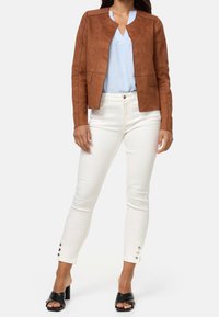 Brun fuskmockajacka med en skräddarsydd design, tillsammans med en ljusblå blus och vita smala jeans med knappdetaljer. Svarta klacksandaler med blockheel.