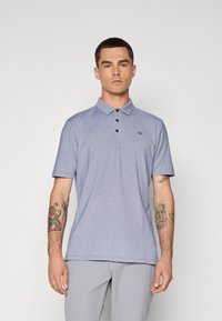 Polo majica s kratkimi rokavi v svetlo modri tkanini, ki ima ovratnik, zapiranje na tri gumbe in majhen logo na prsih.