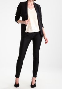 Svart skräddarsydd blazer över en vit paljett-topp, kombinerat med figurnära svarta byxor och svarta högklackade skor, mot en enkel bakgrund.