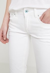Los pantalones vaqueros de mezclilla blanca tienen un diseño ajustado, estilo de cinco bolsillos y un botón de metal azul. La tela tiene un acabado suave y ligeramente texturizado.
