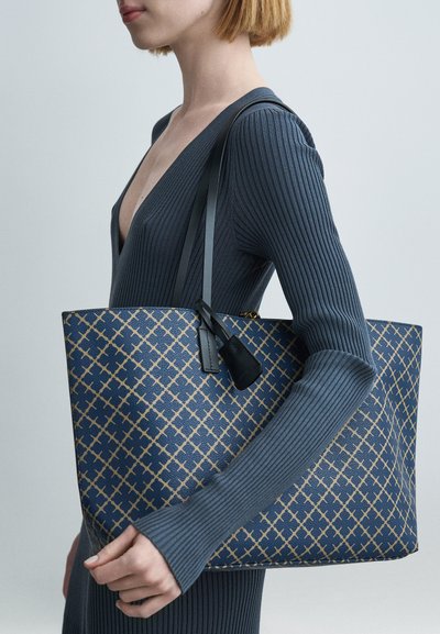 Sac cabas bleu marine avec un motif géométrique doré. Matériau texturé, forme plate et poignées noires contrastantes avec une petite étiquette accent.