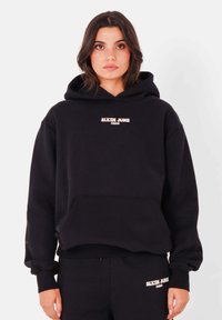 Zwarte hoodie met een voorkentas, voorzien van "SIXTH JUNE PARIS" in witte borduursel. Zachte textuur, oversized pasvorm, ribgebreide manchetten en zoom.