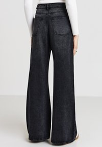 Pantalon en denim noir à jambes évasées, présentant une taille haute, des poches arrière et un tissu légèrement texturé avec un léger éclat.