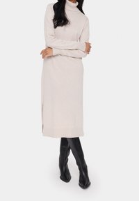 Beige dlouhý rukáv turtleneck šaty s postranními rozparky, vyrobené z měkké, strukturované látky. Nošeny s úzkými černými kozačkami po kolena.