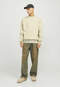 Sudadera beige con hombros caídos sobre una camiseta de tie-dye clara, combinada con unos pantalones vaqueros holgados de color verdoso-marrón y zapatos tipo slip-on beige. Gorra negra.