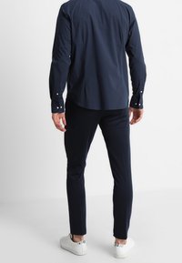 Camisa azul marino de botones con mangas largas, combinada con pantalones ajustados azul marino y zapatillas blancas con detalles estampados, vista desde atrás.