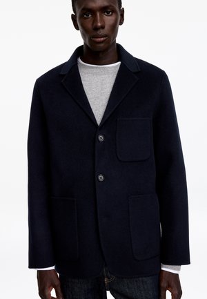 Homme à la peau foncée portant un blazer bleu marine foncé avec trois poches avant par-dessus un pull gris clair sur un fond blanc uni.