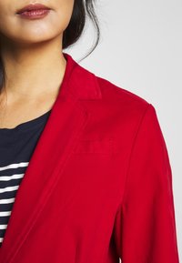 Červený blazer s hladkou texturou, klopovým límcem a jednou náprsní kapsou. Nesený přes námořnický pruhovaný košili.