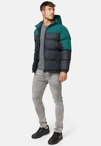 Kurtka puchowa w ciemnoszarym kolorze z akcentami w odcieniu teal, z kapturem, zapięciem na zamek i pikowanym wzorem. Noszona z szarymi dżinsami i czarnymi sneakersami.