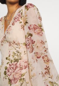 Vestido floral con mangas largas, translúcidas y abullonadas. Tejido de color beige claro adornado con patrones de flores en rosa y verde. Diseño en forma de V.