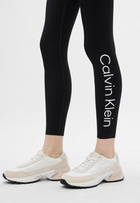 Fekete leggings, fehér "Calvin Klein" logóval, amely függőlegesen van nyomtatva; fehér és bézs sportcipőkkel párosítva, amelyek texturált felülettel és gumitalppal rendelkeznek.