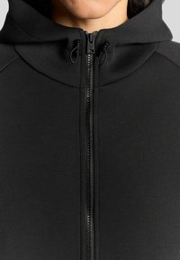 Svart zip-up hoodie med hög krage, med en mjuk tygkvalitet, plastdragkedja och snörningsdetaljer vid huvan.