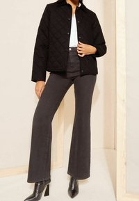 Černá prošívaná bunda se cvoky, kombinovaná se šedými zvonovými kalhotami a černými kotníkovými botami, na neutrálním pozadí.