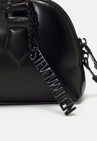 Sac à main en simili cuir noir, de forme arrondie, avec une anse en chaîne et un tirette de fermeture à glissière de marque affichant clairement le texte "STEVE MADDEN".