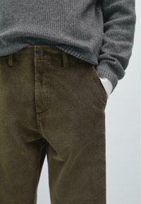 Person iført olivengrønne corduroybukser og en grå ribbestrikket sweater, med den ene hånd i lommen mod en ensfarvet baggrund.