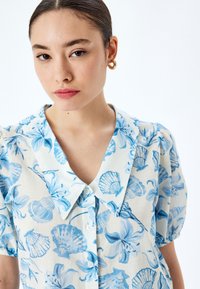 Leichte, kurzärmelige Bluse aus hellem Stoff mit blauen Blumen- und Muschelmustern. Verfügt über einen großen Kragen und Knöpfe auf der Vorderseite.