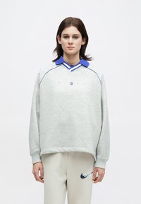 Νέος άνθρωπος που φοράει ένα ανοιχτό γκρι φούτερ με μπλε γιακά και μπεζ παντελόνι που διαθέτει το σκούρο μπλε λογότυπο της Nike, στέκεται μπροστά σε ουδέτερο φόντο.