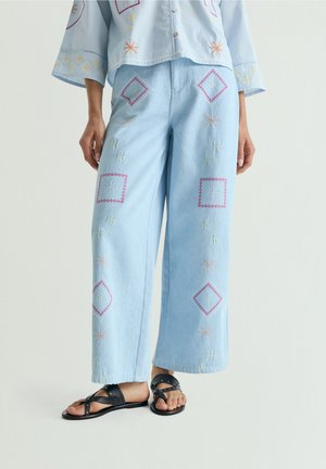 Pantalones azul claro de pierna ancha con bordado geométrico colorido, combinados con una camisa bordada a juego y sandalias negras con tachuelas.