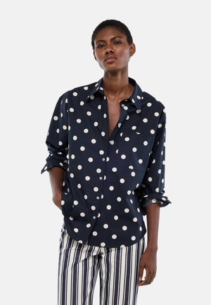 Femme portant une chemise bleu marine à pois avec manches retroussées et un pantalon rayé verticalement bleu marine et blanc, debout avec une main dans la poche.