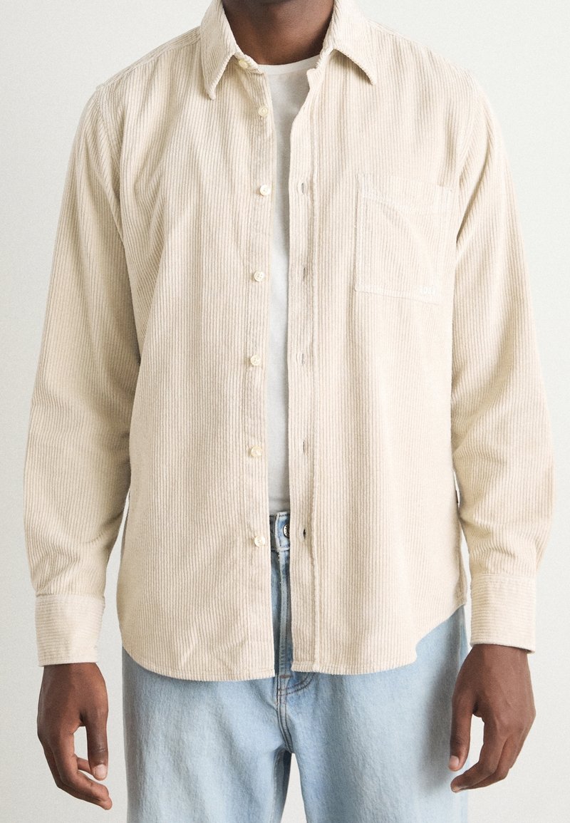 Homme portant une chemise beige en velours côtelé à boutons sur un t-shirt blanc et un jean bleu clair, visible des épaules jusqu'aux hanches.