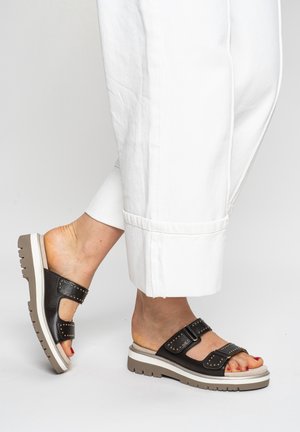 Füße in schwarzen Ledersandalen mit zwei mit Nieten besetzten Riemen und weißer Sohle, kombiniert mit weißen Caprihosen.