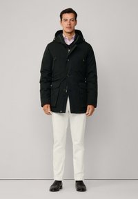 Muškarac nosi crnu zimska jaknu s kapuljačom preko prugaste košulje i crnog pulovera, bijelih hlača i crnih poslovnih cipela na neutralnoj pozadini.