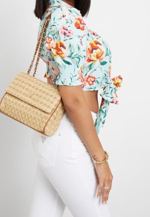 Kobieta w kwiecistej krótkiej bluzce i białych spodniach, niosąca beżową teksturowaną torebkę na ramię z łańcuszkowym paskiem na białym tle.