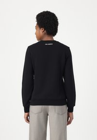 Črn pulover s dolgimi rokavi, okroglico na vratu, rebrastimi manšetami in robom. Na hrbtni strani je vidna bela ikona Karl Lagerfeld.