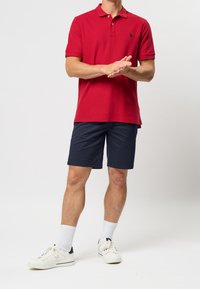 Czerwona koszulka polo z krótkimi rękawami i kołnierzykiem, zestawiona z granatowymi szortami. Dopełnieniem stroju są białe trampki i skarpetki.