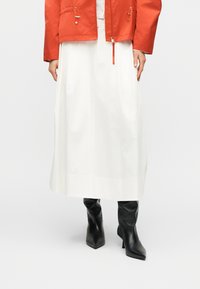 Jupe midi blanche associée à des bottines à talons noires à bout pointu et une veste orange avec détails zippés et cordon de serrage.