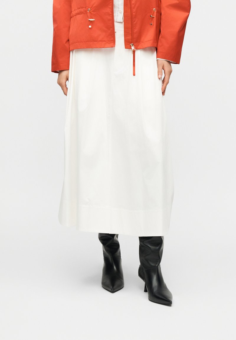 Jupe midi blanche associée à des bottines à talons noires à bout pointu et une veste orange avec détails zippés et cordon de serrage.