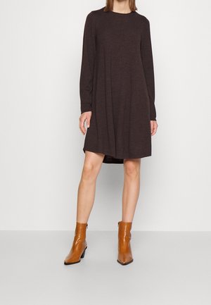 Mujer con un vestido de manga larga color marrón oscuro hasta la rodilla, combinado con botines color canela, delante de un fondo liso claro.