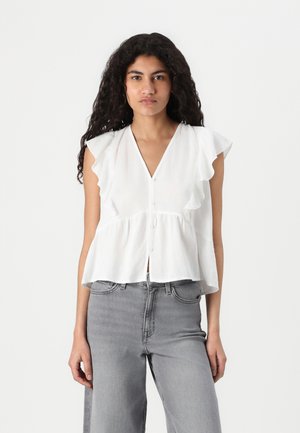 Blouse peplum blanche avec des manches à volants, décolleté en V et fermeture à boutons sur le devant, associée à un jean taille haute gris. Tissu léger et texturé.