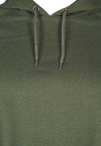 Gros plan sur le tissu d'un sweat à capuche vert olive avec deux cordons passés à travers des œillets métalliques au niveau de l'encolure.