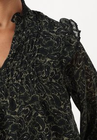 Mørkegrønn bluse i lett, teksturert stoff. Har et paisley-mønster, rynket halslinning og volanger på skuldrene.