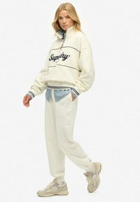 Crème sweatshirt met zwarte letters, gestreepte mouwen en een rits-kraag, gecombineerd met bijpassende crème broeken en beige sneakers. Lichtblauw binnenhemd.