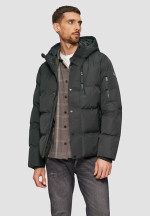 Dunkelgrüne Pustejacke mit Kapuze, ausgestattet mit Reißverschlusstaschen und einer glatten Textur. Getragen über einem karierten Hemd und verblassten Jeans.