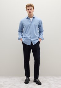 Camicia azzurro chiaro con bottoni e tessuto liscio, abbinata a pantaloni di velluto a coste scuri. Modello in piedi con le mani nelle tasche, indossa scarpe nere.