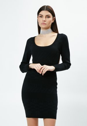 Abito bodycon nero con maniche lunghe, caratterizzato da una texture a coste e la scritta "INFLUENCER". Tessuto liscio e scollatura a barchetta. Accessorio choker argentato.
