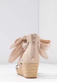 Beżowe sandały na koturnie z plecioną rope podeszwą, z tylnym zamkiem błyskawicznym oraz dużą beżową kokardą z materiału zawiązaną wokół kostki na białym tle.