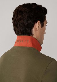 Muži s tmavými vlasmi je oblečený v olivovozelenom saku s vyvýšeným oranžovým golierom vyšitým nápisom "HACKETT," otočený chrbtom na neutrálne pozadí.