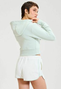 Sudadera corta verde claro con cordón y puños de canalé, combinada con pantalones cortos blancos texturizados que presentan un borde en tonos pastel y slit laterales.