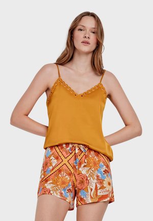 Vrouw met lang haar die een mosterdgele top en korte broek met bloemmotief in oranje, blauw en wit draagt, staand met handen achter de rug.