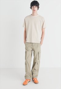 Beige T-Shirt mit kurzen Ärmeln kombiniert mit hellkhakifarbenden Cargo-Hosen mit mehreren Taschen. Orangefarbene Sneakers runden das Outfit ab.