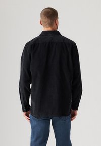 Svart kordflordsskjorta med långa ärmar, knäppningar vid ärmsluten och en ryggparti detaljer, i kombination med blå jeans. Texturen ser mjuk och ribbad ut.
