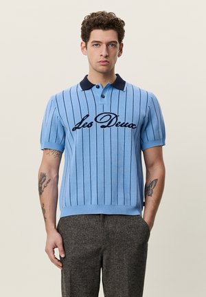 Jeune homme aux cheveux bouclés porte un pull polo à manches courtes bleu clair avec des rayures verticales noires et le texte "Les Deux", assorti à un pantalon foncé.