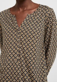 Bluse mit Knopfleiste auf dunklem Hintergrund, mit einem gold- und cremefarbenen geometrischen Muster, leichtem V-Ausschnitt und Textur des Stoffes.